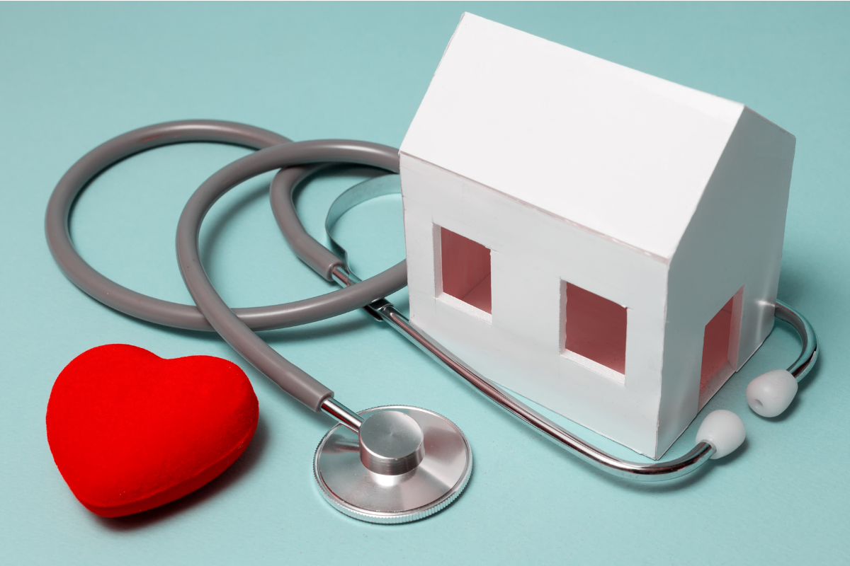 A wooden house is shown with a stethoscope and a red heart, representing the relationship between housing status and health